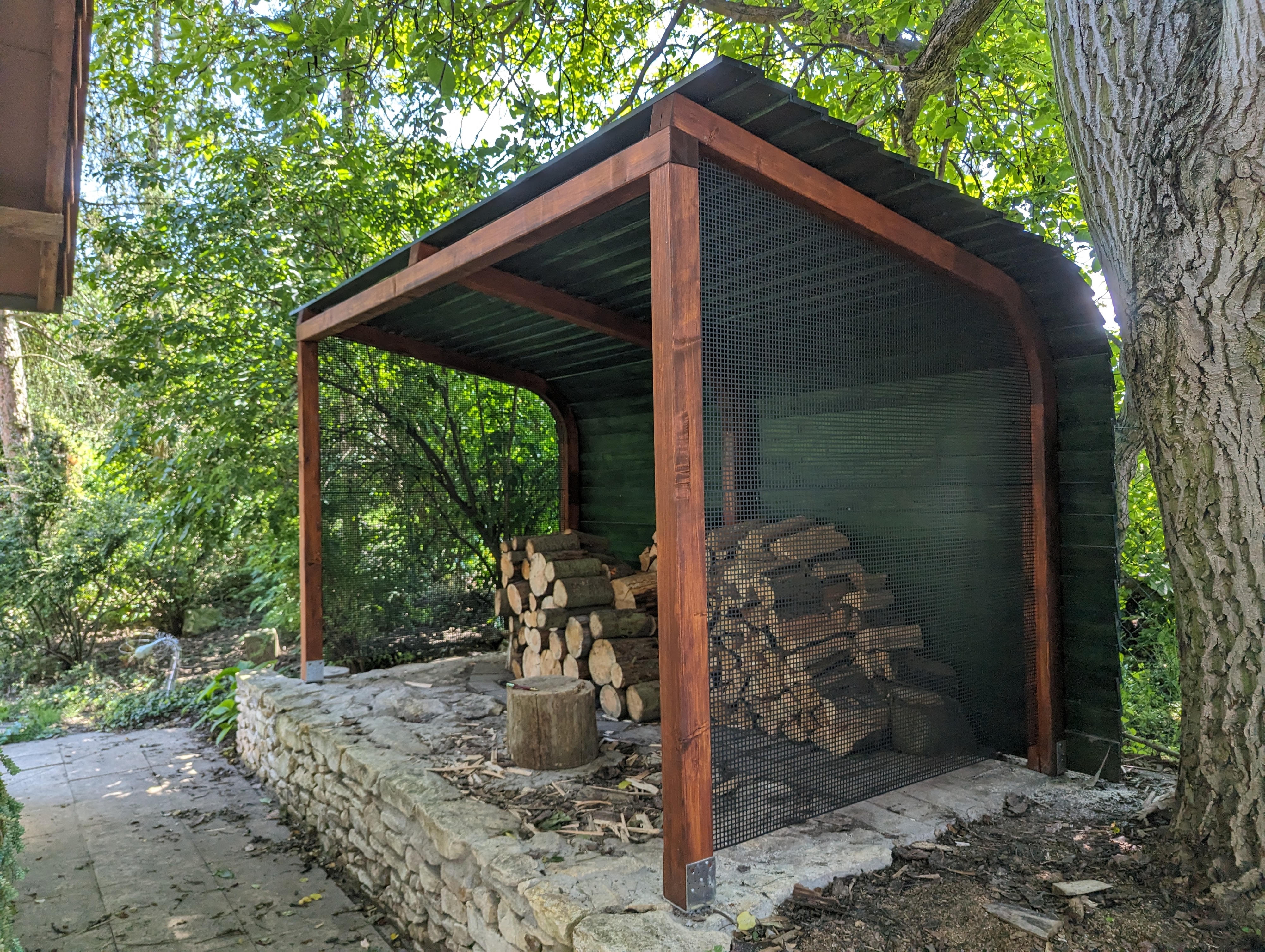Woodgon — woodshed with curved roof, mesh side, and chopping block in a garden