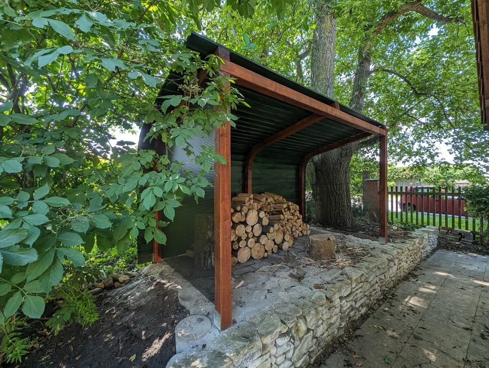 Woodgon — wooden woodshed on a stone wall in a garden with stacked firewood